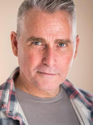 Carl Christopher – The REV Theatre Co. | Broadway in the Finger Lakes Close-up portrait of a middle-aged man with short gray hair, wearing a gray shirt and plaid shirt, looking at the camera.