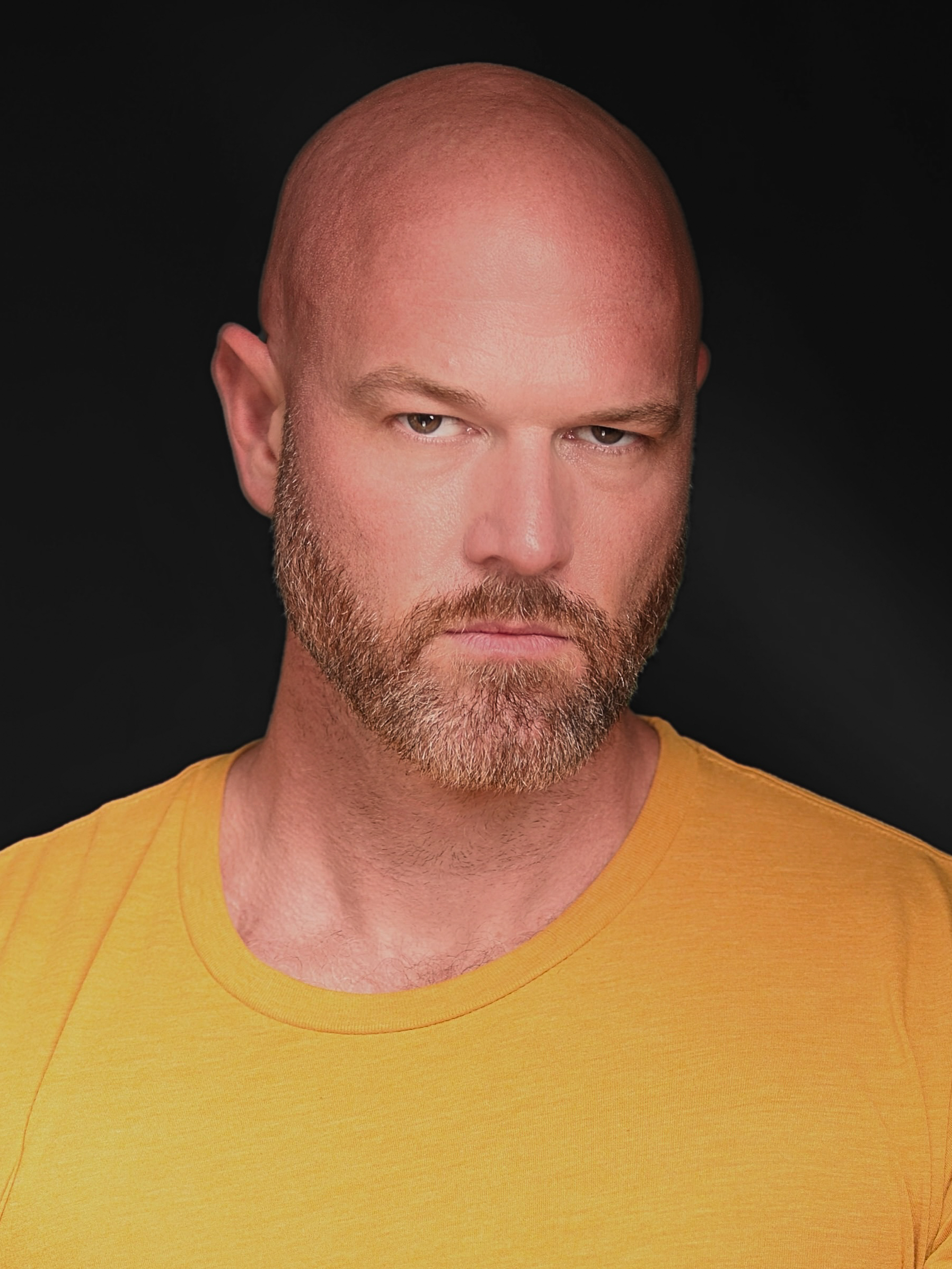 James Zachary – The REV Theatre Co. | Broadway in the Finger Lakes Close-up portrait of a bald man with a beard wearing a yellow shirt against a black background, looking slightly to the side.
