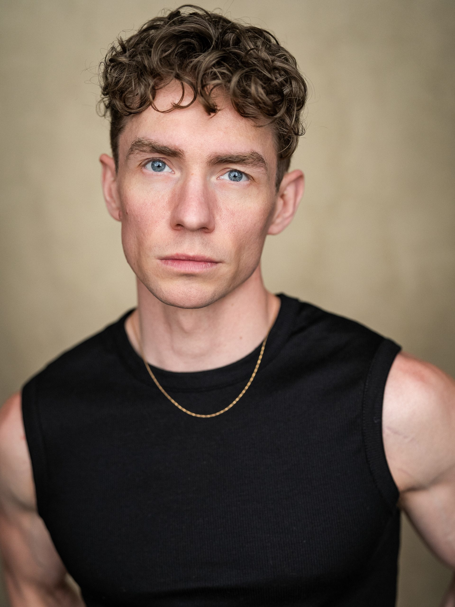Kringer Jeffery – The REV Theatre Co. | Broadway in the Finger Lakes Close-up portrait of a man with blue eyes, wearing a black sleeveless shirt and a thin gold chain against a neutral background.