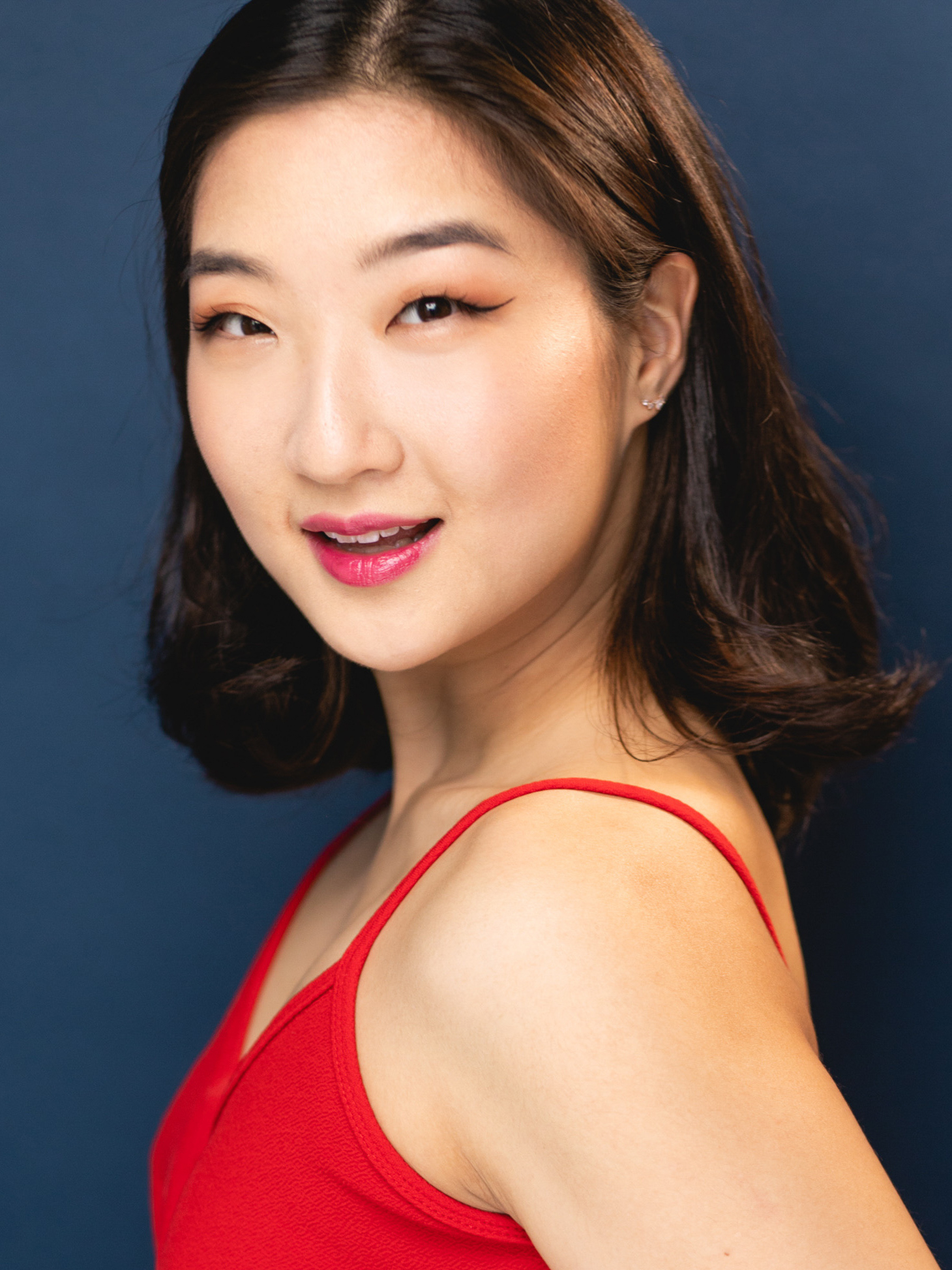 Lee Esther – The REV Theatre Co. | Broadway in the Finger Lakes Portrait of an Asian woman in a red dress against a dark blue background, facing slightly to the left.