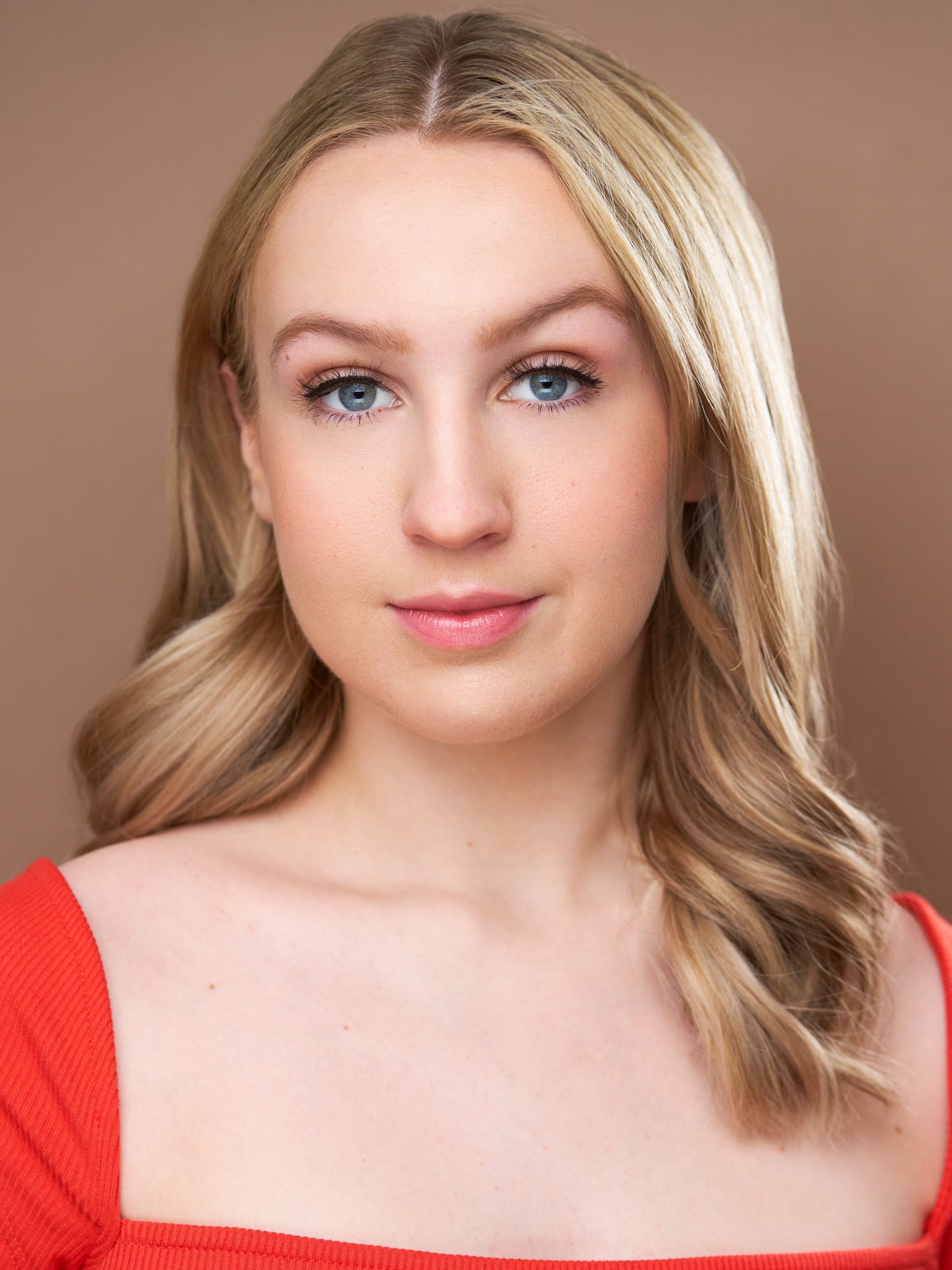 Lentz Maddie – The REV Theatre Co. | Broadway in the Finger Lakes Close-up portrait of a blonde woman with blue eyes wearing a red off-shoulder top, neutral brown backdrop.