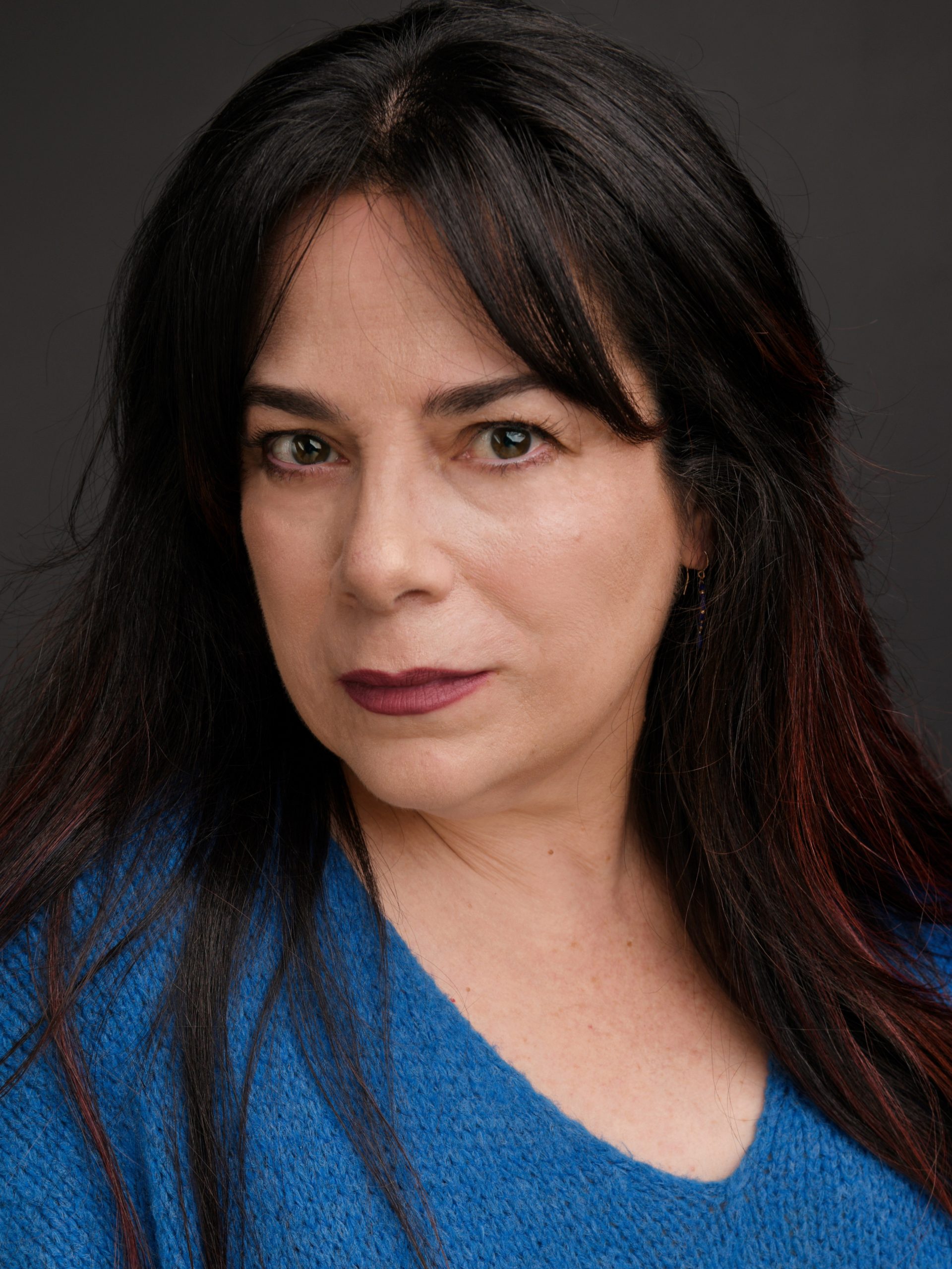 Madeoy Celia – The REV Theatre Co. | Broadway in the Finger Lakes Close-up portrait of a woman with dark hair and brown eyes, wearing a blue knit sweater against a dark background.