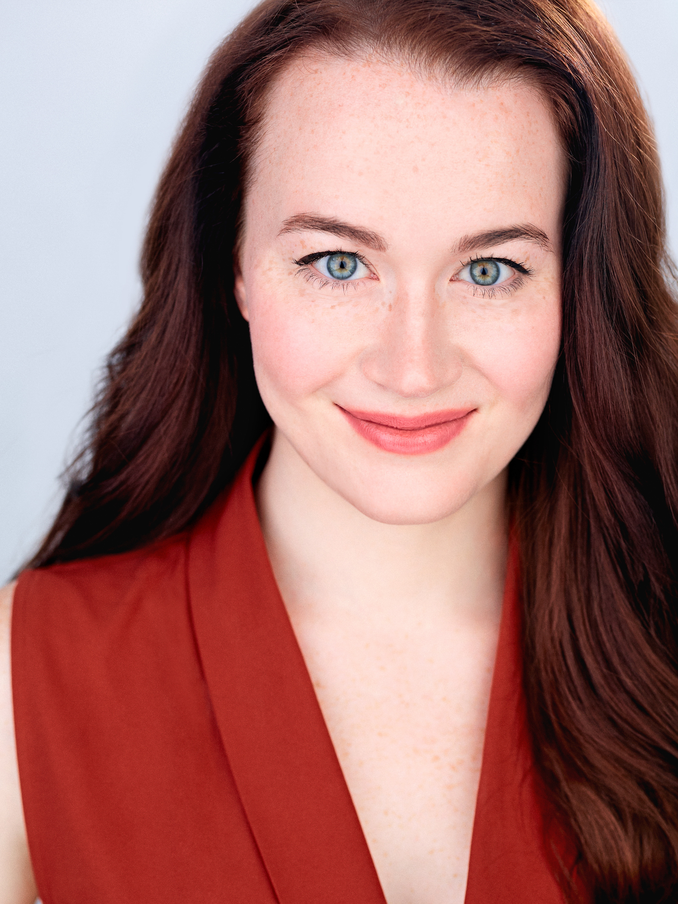 Malaney Mary – The REV Theatre Co. | Broadway in the Finger Lakes Close-up portrait of a woman with blue eyes and red hair wearing a red blouse, smiling at the camera.