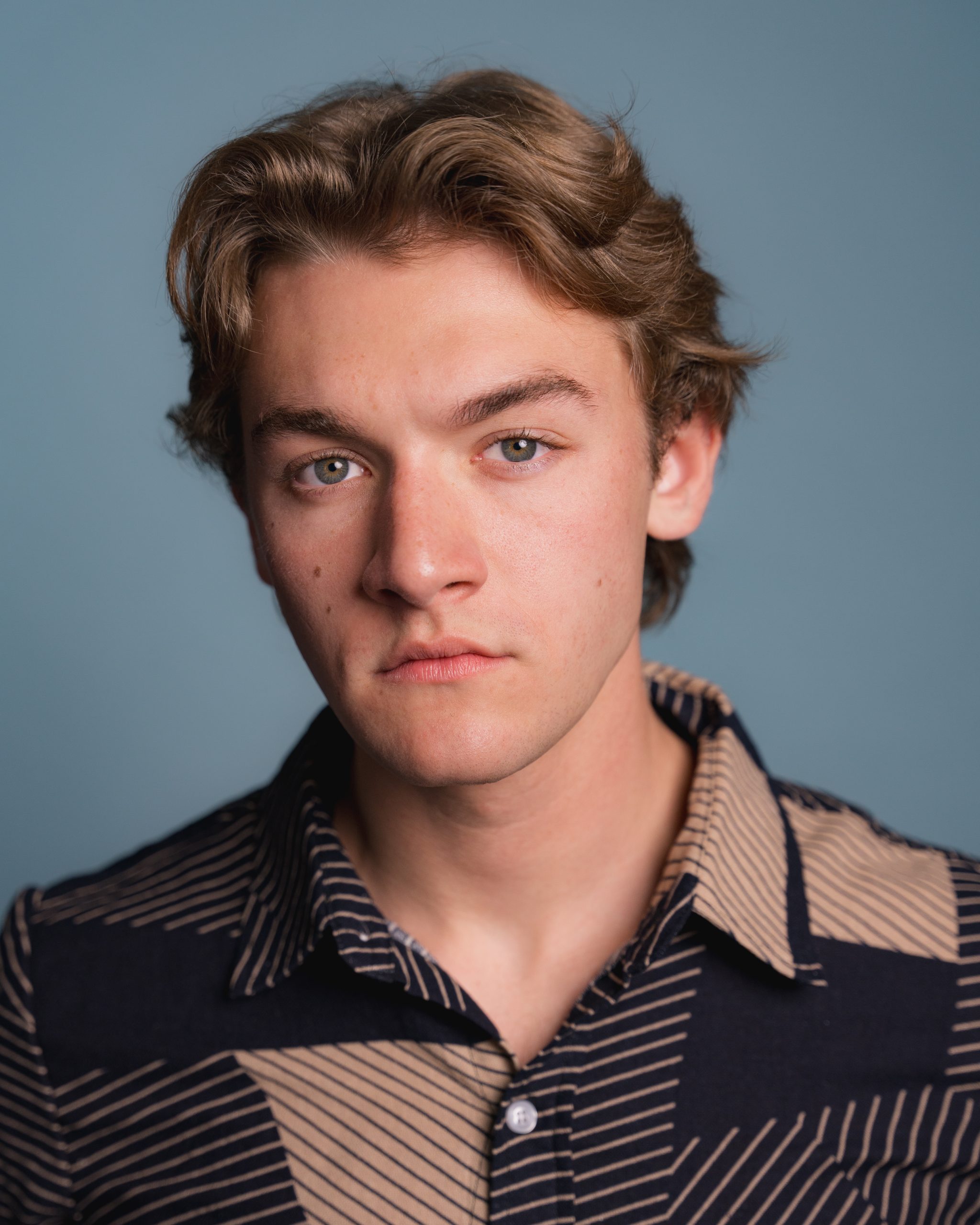 Asa Naigus – The REV Theatre Co. | Broadway in the Finger Lakes Close-up portrait of a young man with light brown hair and blue eyes, wearing a navy striped shirt against a blue backdrop.