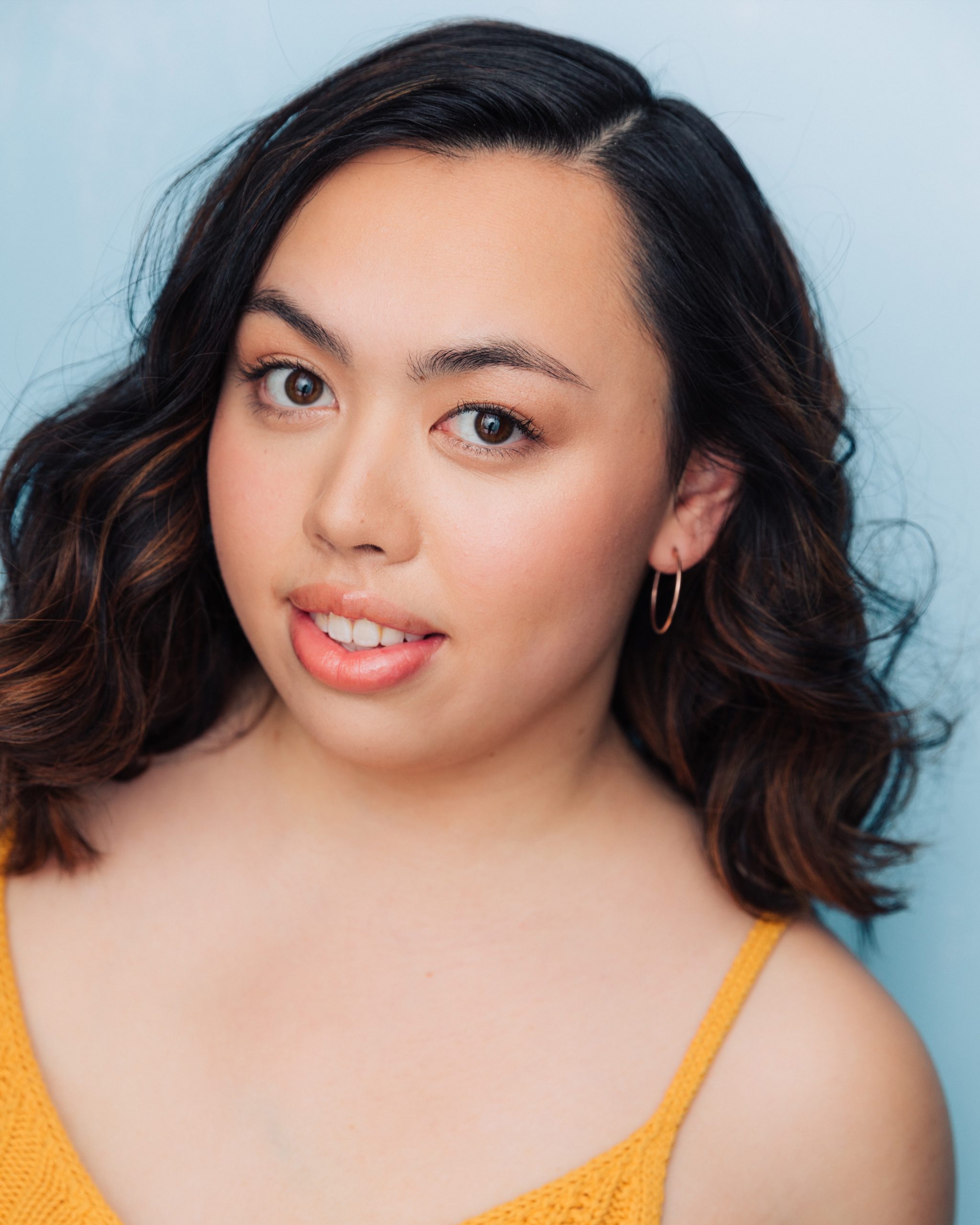 Neuschulz Kimmi – The REV Theatre Co. | Broadway in the Finger Lakes Close-up of a young woman with dark hair and brown eyes, wearing a yellow top against a blue background.