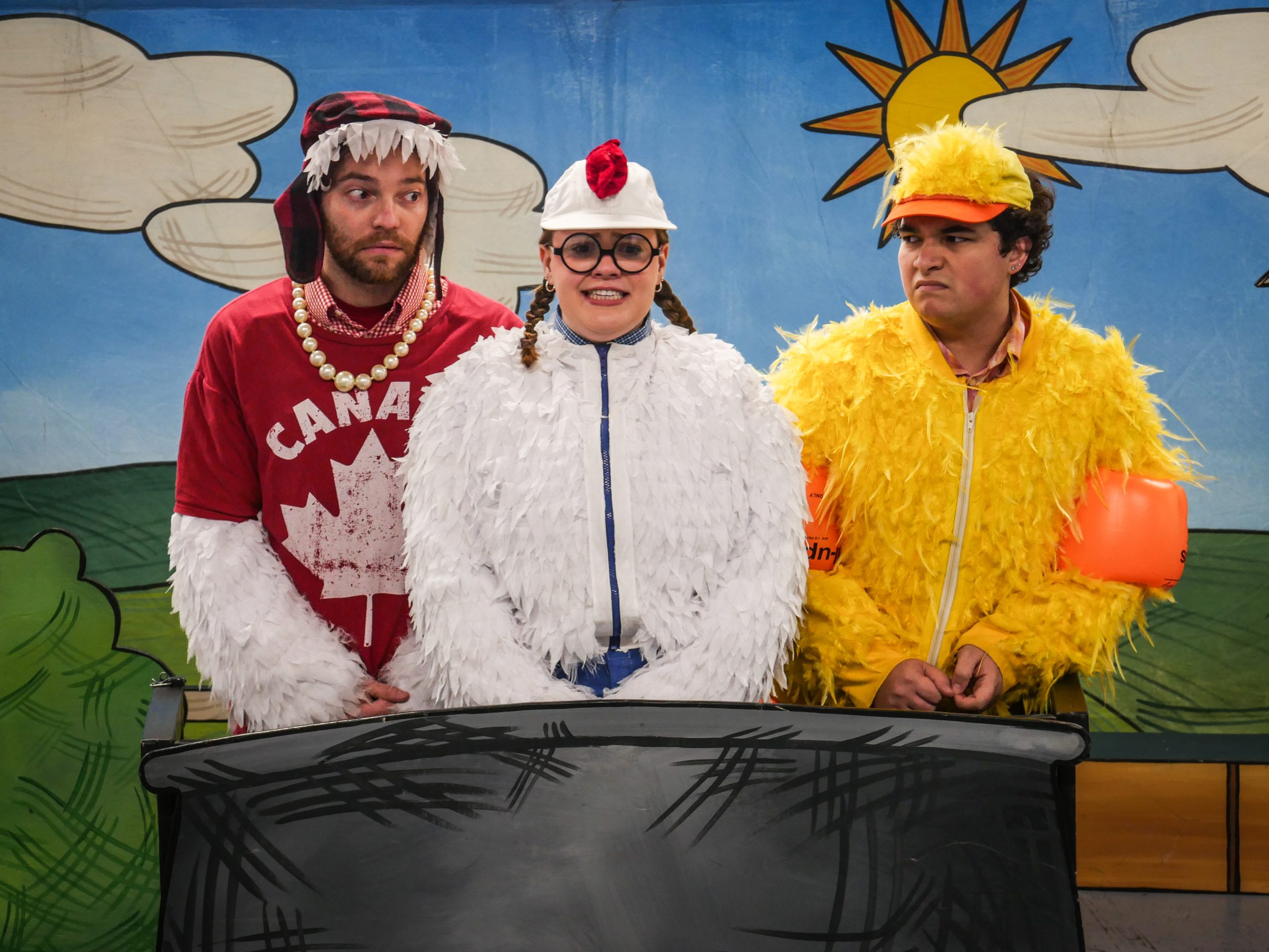 Three performers in colorful costumes on a stage: a man in a red shirt with a pearl necklace, a person in a white chicken costume with glasses, and a person in a yellow chicken costume with an orange visor, standing behind a prop table against a cartoon backdrop.