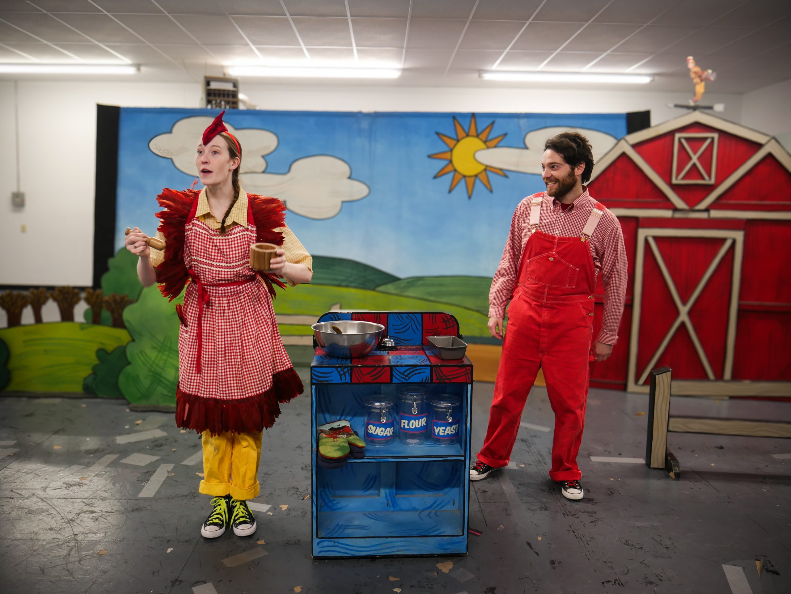 Two performers in a farm-themed stage setting: a woman in a red gingham dress and yellow pants holding wooden mugs, and a man in red overalls smiling, with a painted barn backdrop and props nearby.