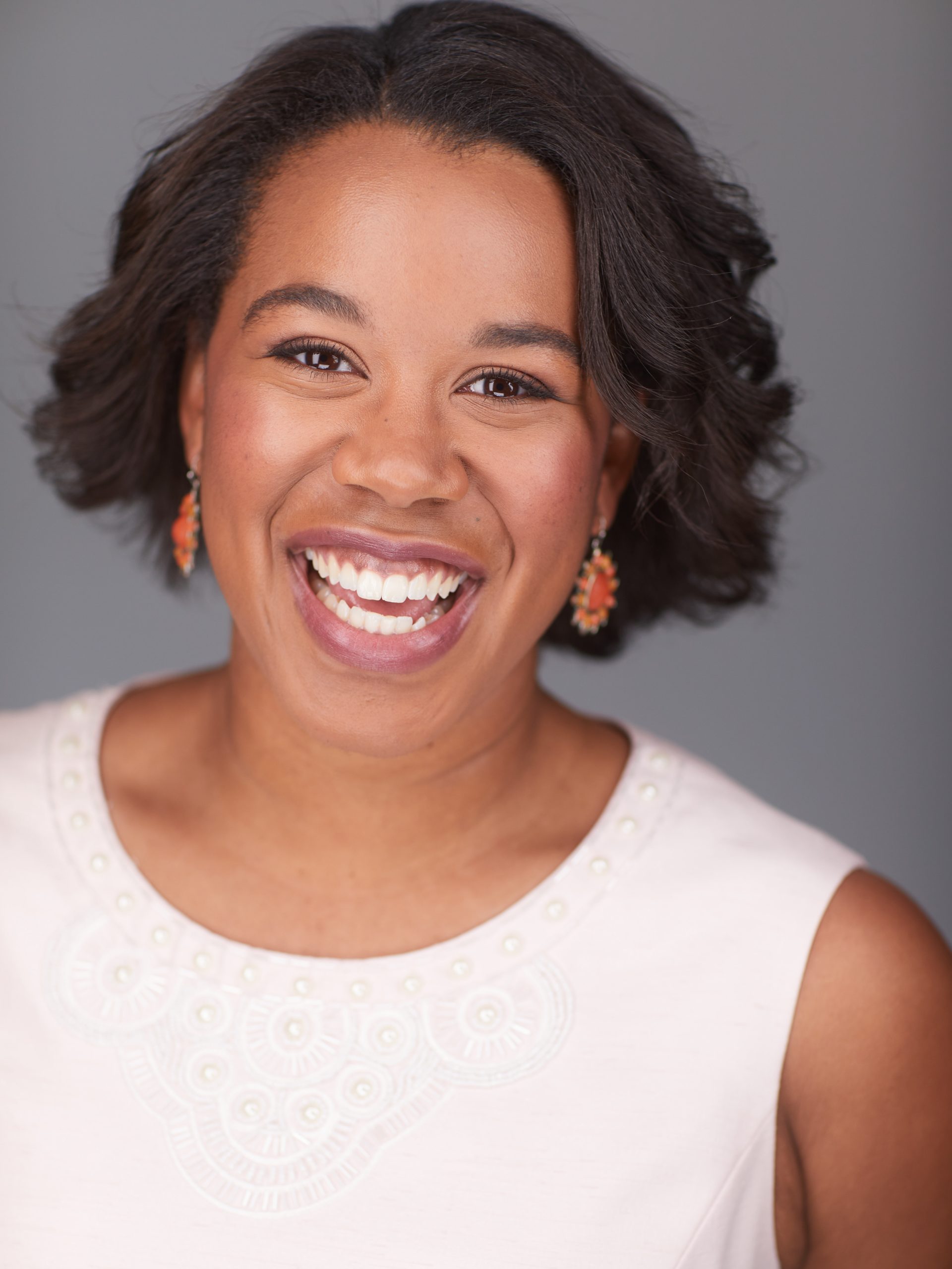 Tagert Alicia Russell – The REV Theatre Co. | Broadway in the Finger Lakes Close-up portrait of a smiling woman with short dark hair, wearing a white embroidered top and orange earrings against a gray background.