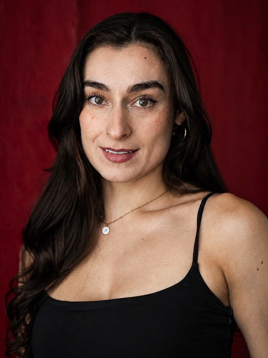 Tia Karaplis Headshot – The REV Theatre Co. | Broadway in the Finger Lakes Portrait of a woman with long dark hair, wearing a thin-strapped black top and a necklace, against a red background.